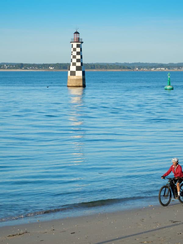 active senior woman cycling with her electric mountain bike along the beach of loctudy in front of the black and white checked lighthouse, brittany, france
